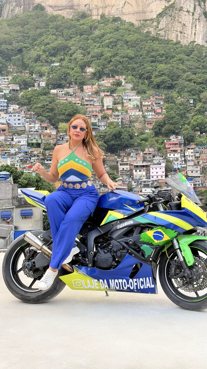 Turista sentada na moto esportiva verde e amarela, com as casas da Rocinha ao fundo.