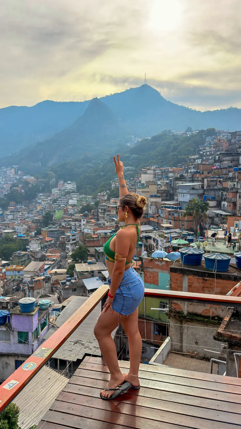 Turista no deck de madeira ao entardecer, com os telhados da Rocinha e as montanhas do Rio ao fundo.
