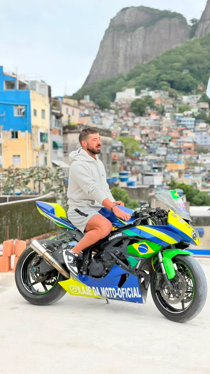 Turista na moto ao lado da bandeira do Brasil, com a Pedra da Gávea ao fundo.