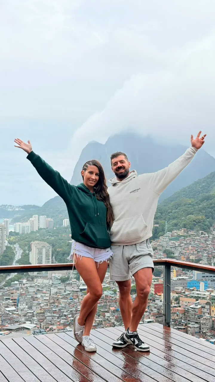 Casal de turistas de braços abertos no deck de madeira, com a Rocinha atrás.