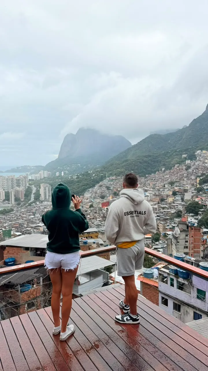 Turista de costas no deck, observando a comunidade e a Pedra da Gávea entre as nuvens.