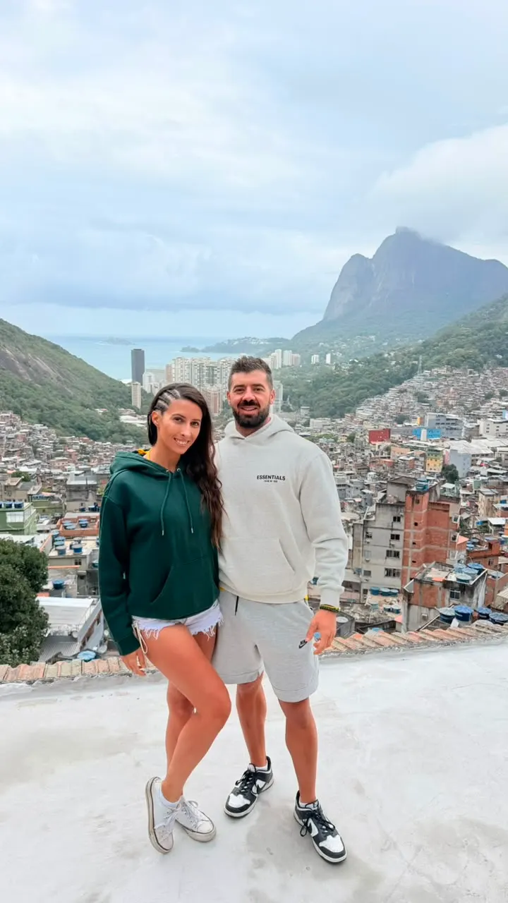 Casal de turistas no mirante, com a comunidade e a Pedra da Gávea ao fundo.